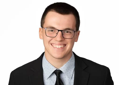 Professional young man wearing glasses, a suit, and a tie, smiling at the camera against a white background.