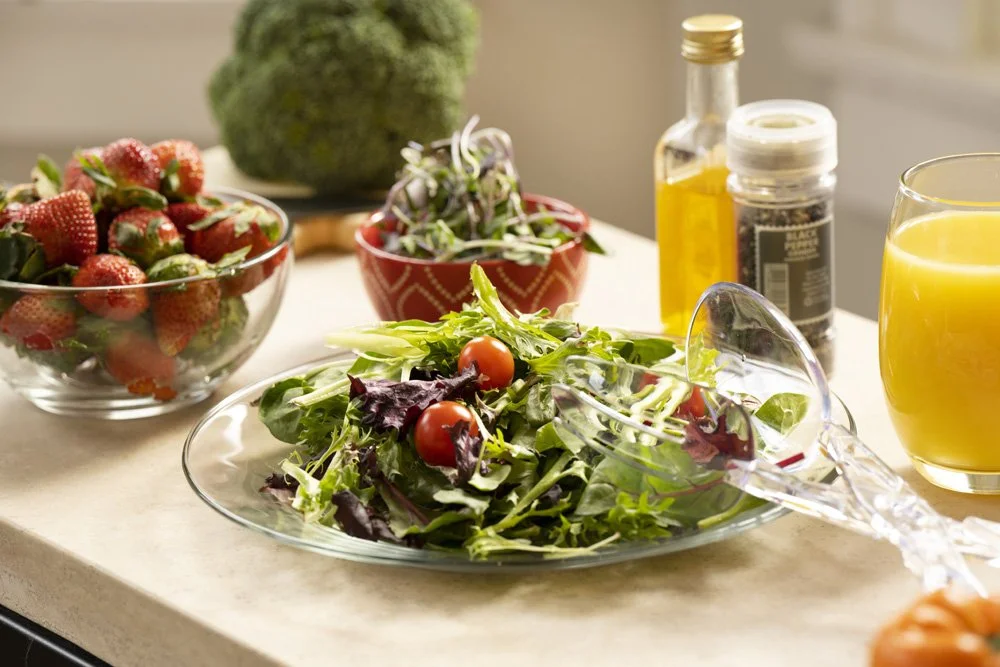 Fresh salad with cherry tomatoes, mixed greens, and purple lettuce on a glass plate, with berries, broccoli, salad toppings, a glass of orange juice, and salad dressing bottles in the background on a kitchen counter.