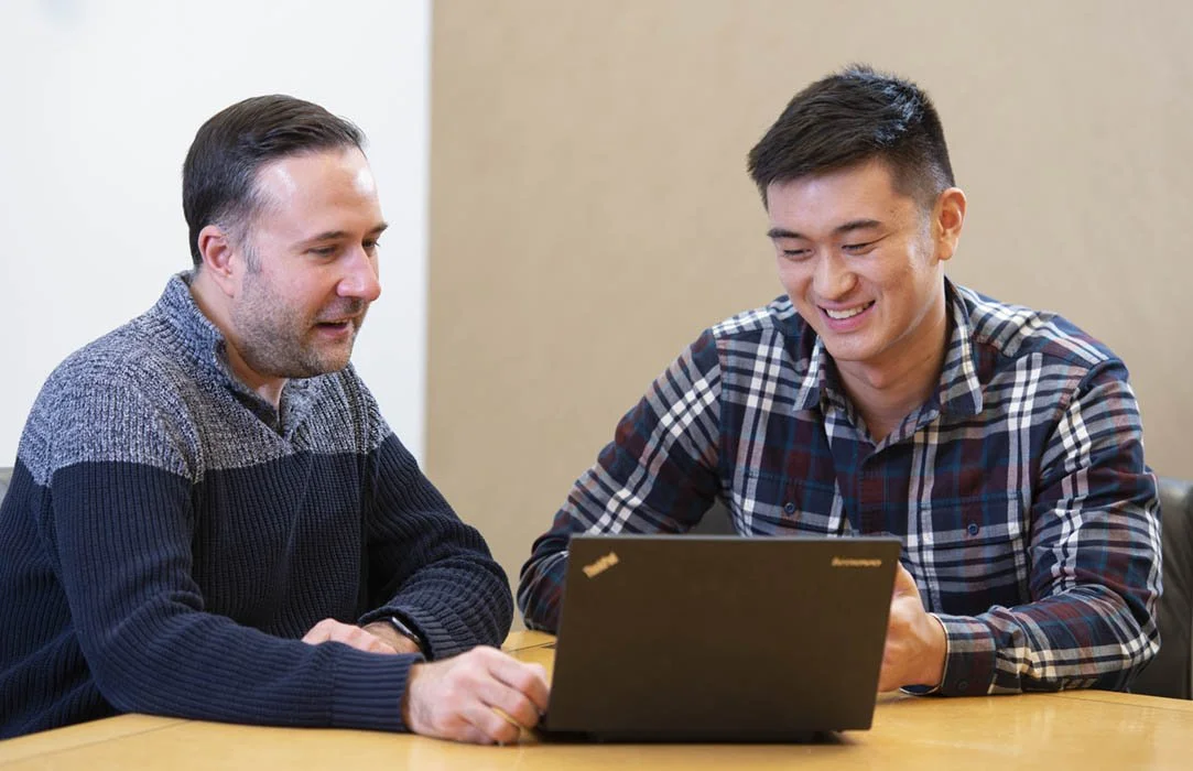 Two men sitting at a table looking at a laptop and smiling in an office taken by a corporate photographer.