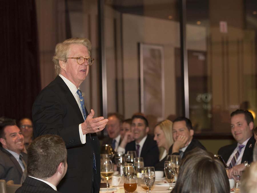 A man with glasses and light-colored hair speaking at a dinner event with several seated attendees laughing and listening.