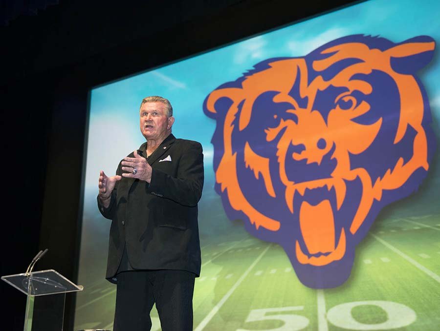 A man in a black suit giving a presentation on a stage with a large screen behind him displaying a stylized tiger head logo.
