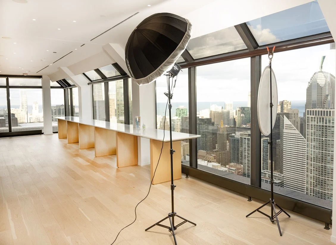 Interior of a modern high-rise space with large windows, photography lighting equipment including a softbox and diffuser, and a long white table, with a city skyline view in the background.