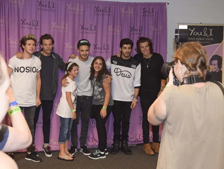 Group of seven people, including five young men and two young girls, posing in front of a purple backdrop with 'You & I' and 'One Direction' logos. A woman with a camera is taking their photo.