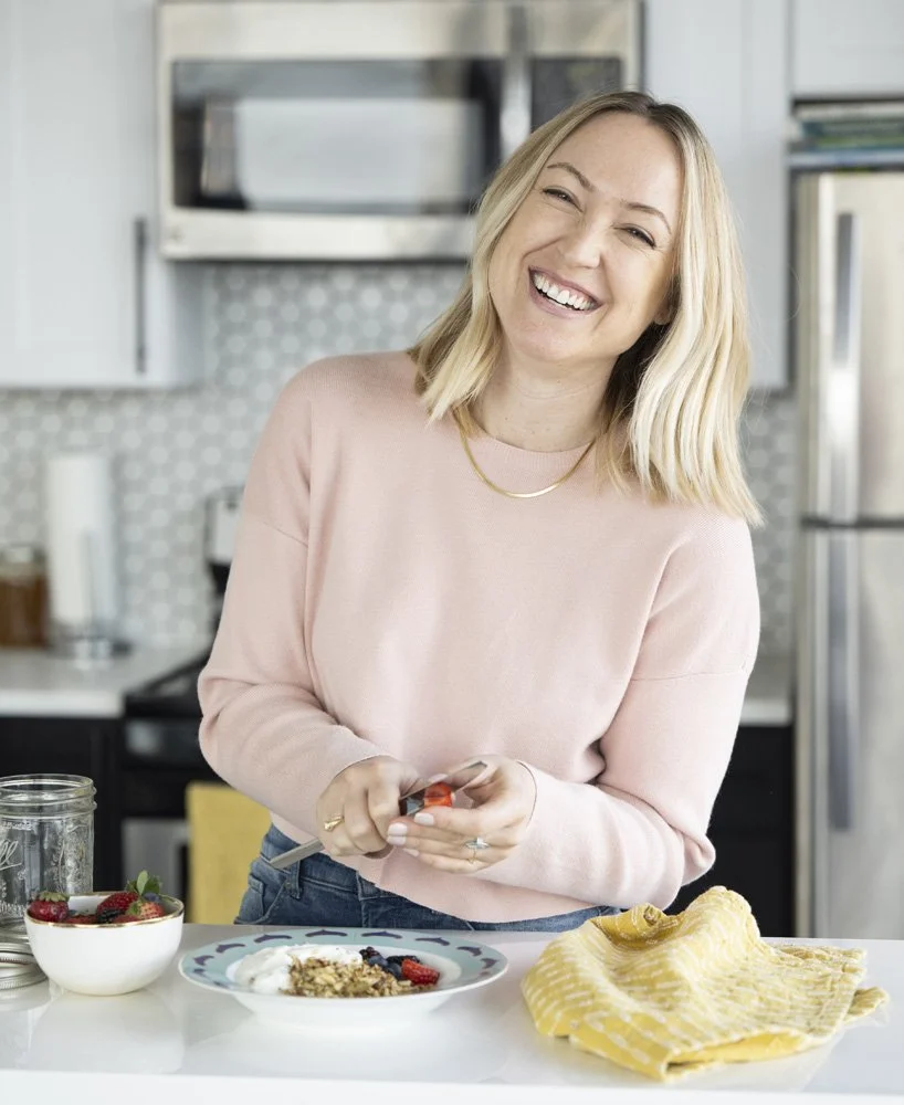 A nutritionist photo shoot smiling in a kitchen, holding a jar opener over a jar, with a bowl of strawberries, a plate of yogurt with granola and berries, and a yellow dish towel on the counter. A personal branding photography session