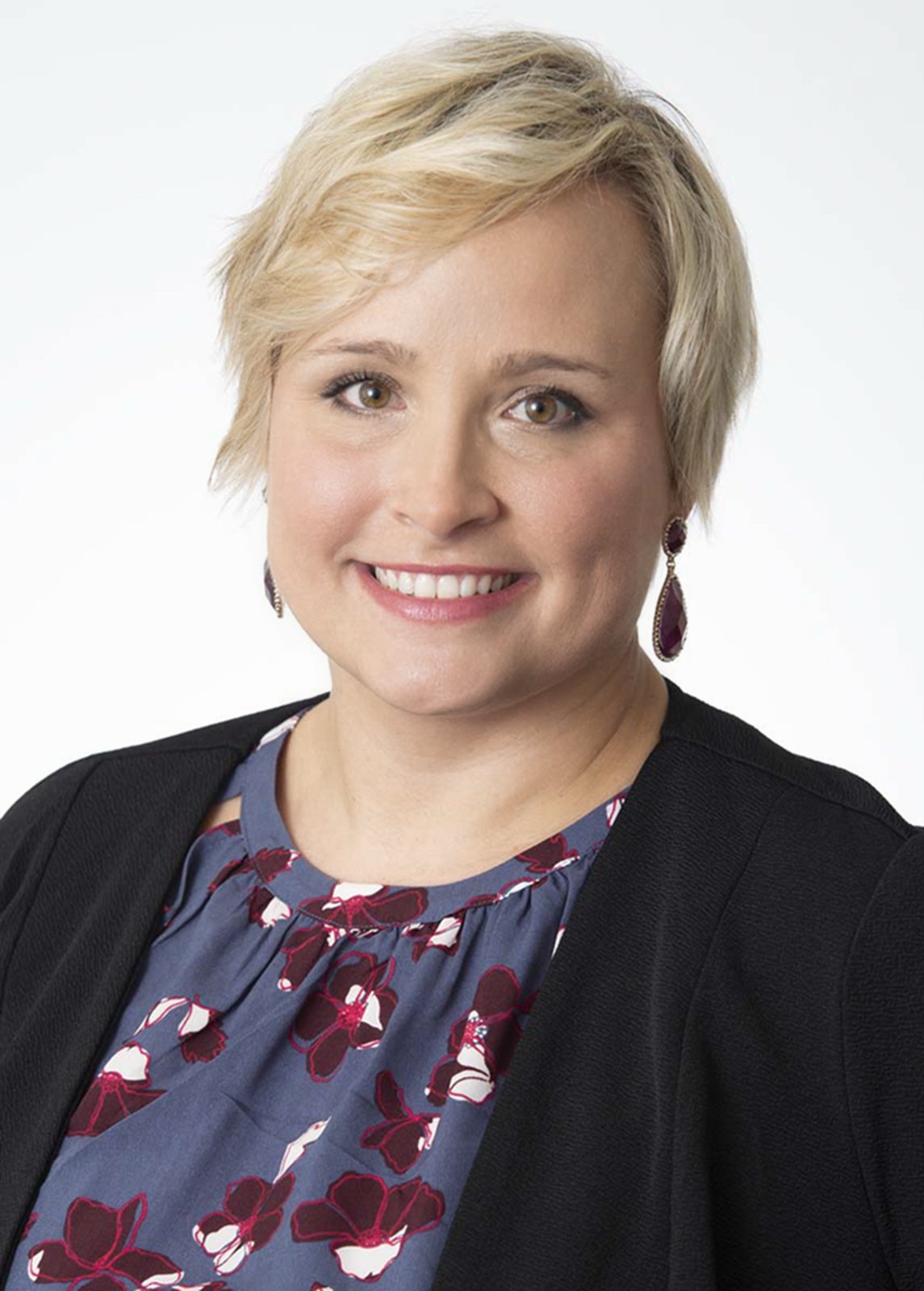 A woman with short blonde hair, wearing a black blazer over a floral dress with red and white flowers, and purple earrings.