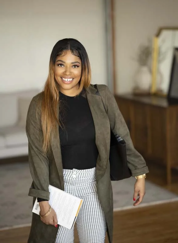 A young woman with long hair is smiling and looking at the camera in an indoor setting. She is holding a notebook in her left hand and wearing a dark top, checked pants, and a green blazer. There is a sofa, a wooden dresser, a mirror, and a vase with