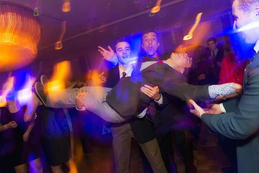 Group of people at a party, lifting and carrying a man horizontally, with colorful, blurred lights in the background.