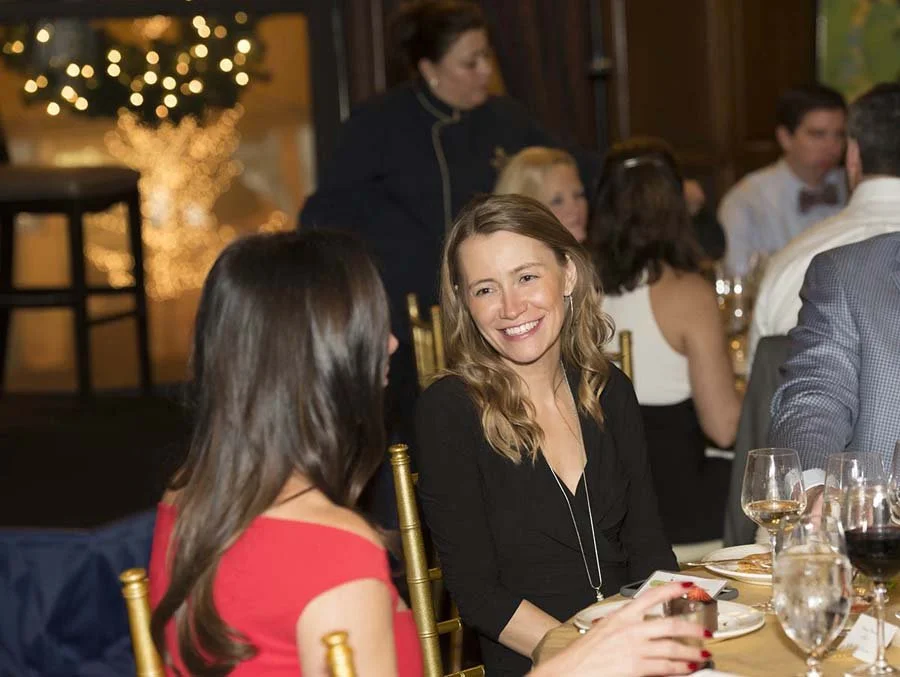 Women sitting at a banquet table, engaged in conversation, smiling, with a decorated Christmas tree in the background.