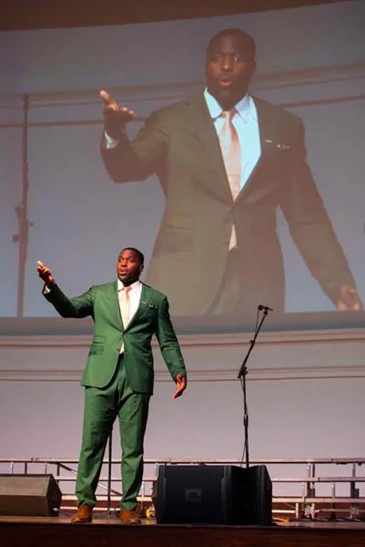 A man in a green suit is speaking on stage, with a large screen behind him displaying a larger image of himself.