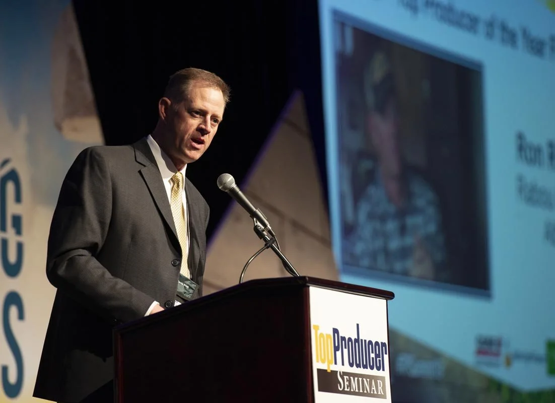 Man in a suit speaking at a podium during a seminar, with a large screen displaying a presentation behind him.