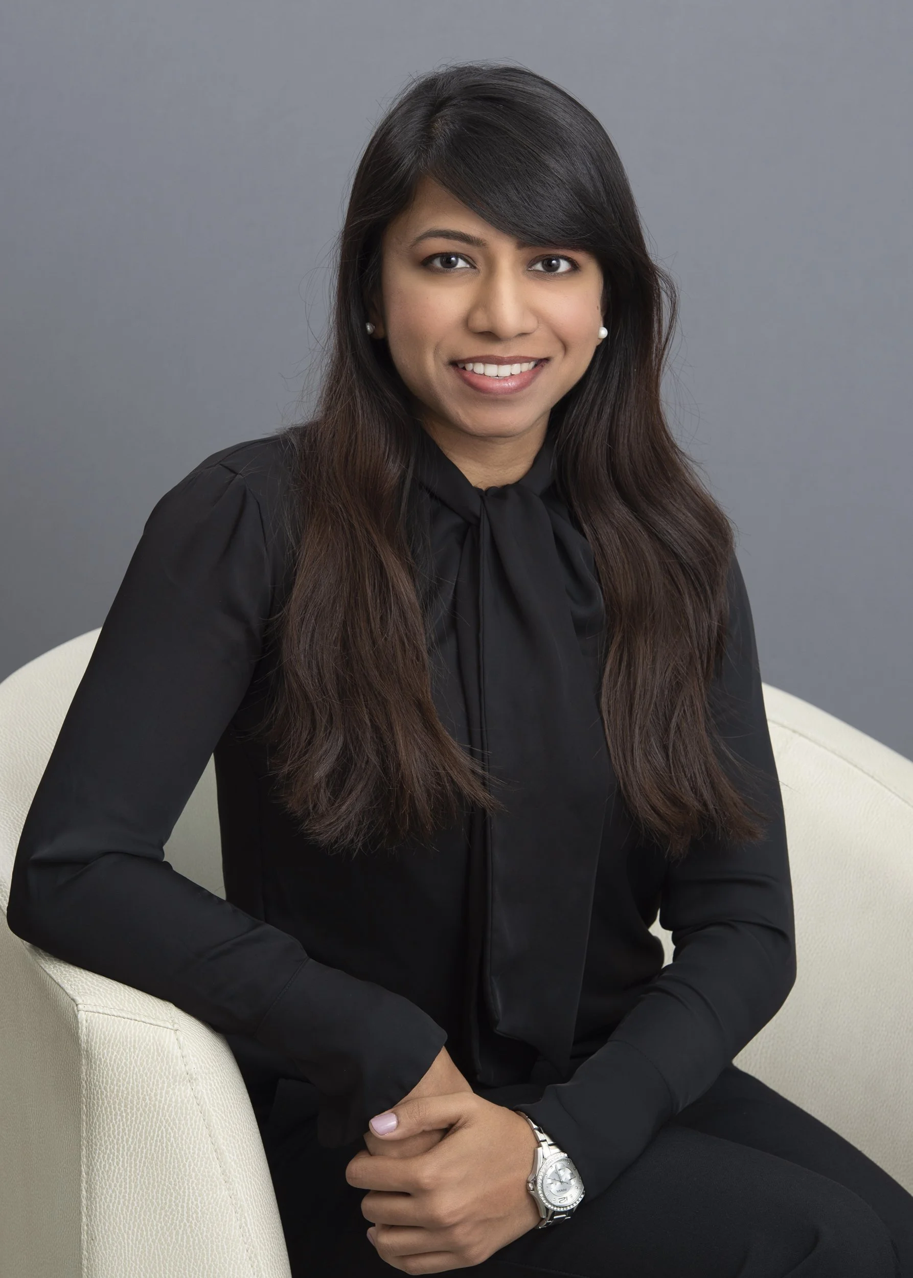 Professional portrait of woman with long dark hair wearing a black blouse, seated on a white chair against a grey studio background, smiling and wearing a silver watch.