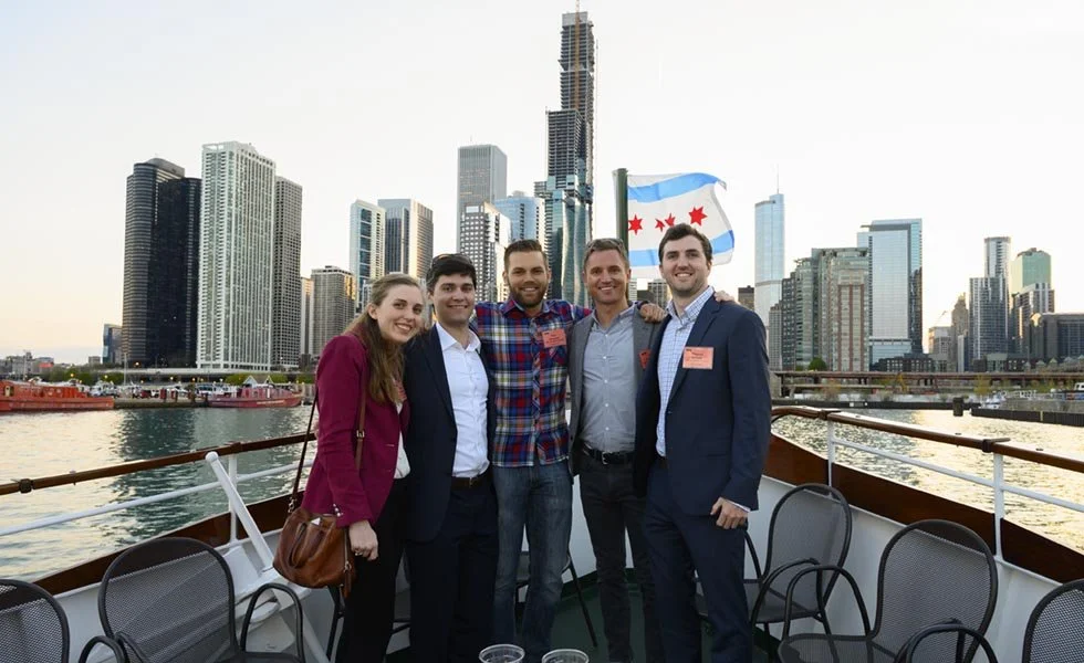 Group of five people standing on a boat with the Chicago skyline in the background, including a flag with blue and white stripes and red stars.