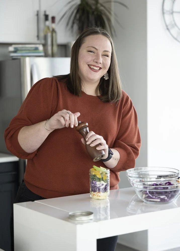 A nutritionist smiling while seasoning a layered salad in a glass jar, with bowls of purple cabbage on the white kitchen counter taken by a Chicago photographer.