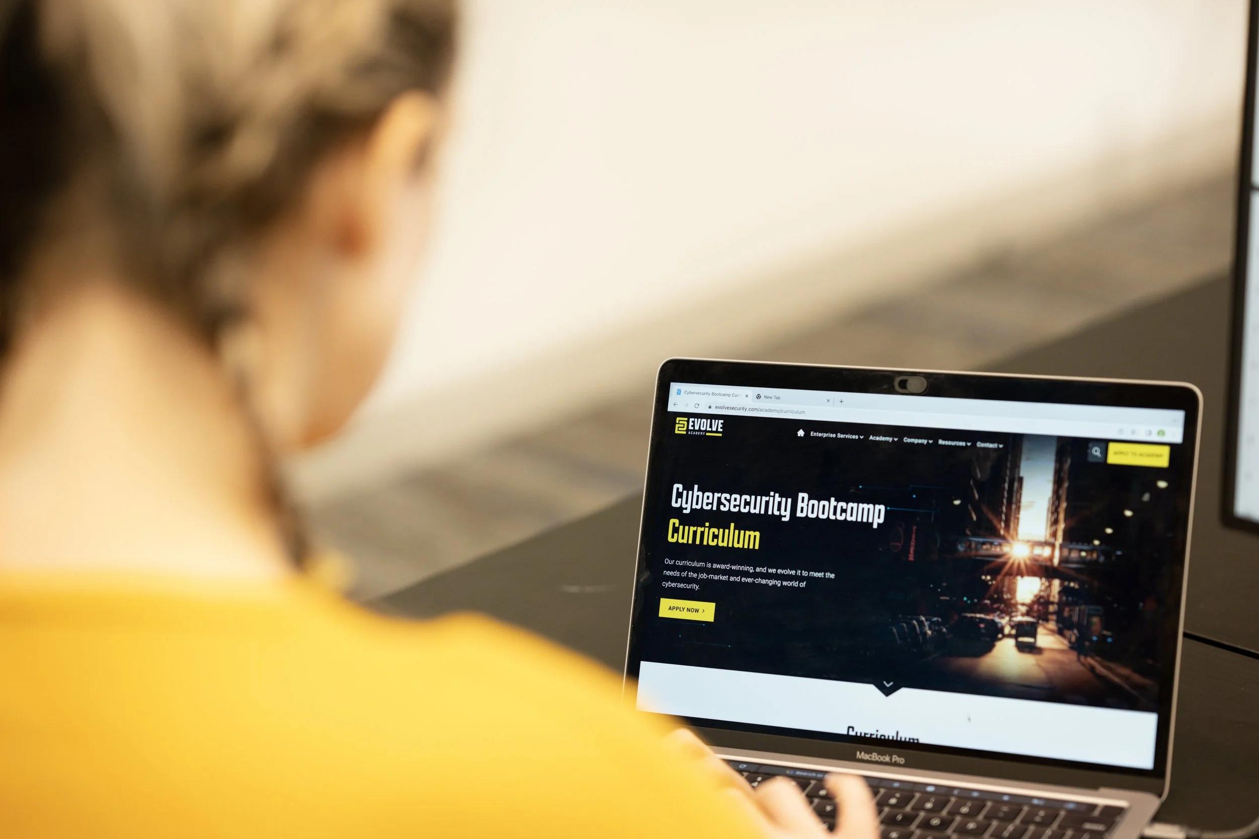 Person wearing yellow shirt using a laptop to view a cybersecurity bootcamp webpage titled 'Cybersecurity Bootcamp Curriculum'.