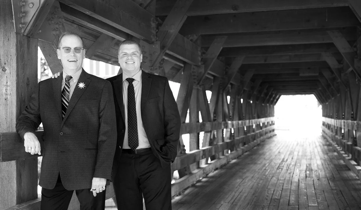Two men in suits standing under a covered wooden bridge, smiling at the camera.