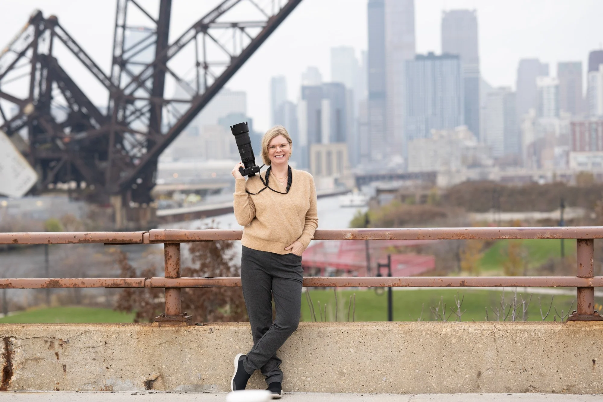 A woman standing on a bridge holding a camera, with a city skyline and a bridge in the background.