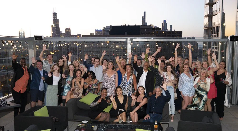 A corporate party celebrating on a rooftop terrace at sunset, many with their hands raised and smiling.