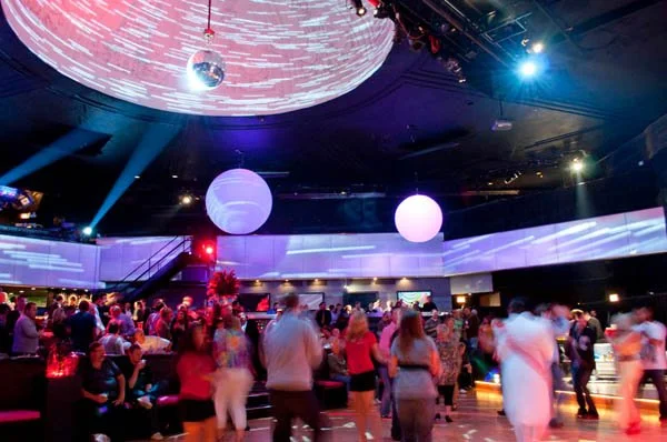 People dancing and socializing at a nightclub with colorful lighting and a disco ball hanging from the ceiling.