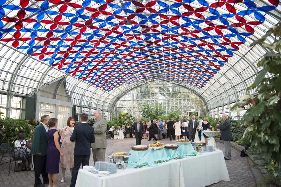 People at a social event in a glass greenhouse decorated with red and blue umbrellas hanging from the ceiling.