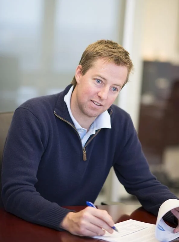 A man with light brown hair and blue eyes sitting at a desk, writing on papers with a pen in his right hand, wearing a navy blue sweater and a light blue collared shirt, in an office setting.
