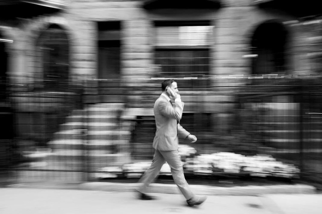A man in a suit walking on the sidewalk while talking on his cellphone in front of a building.