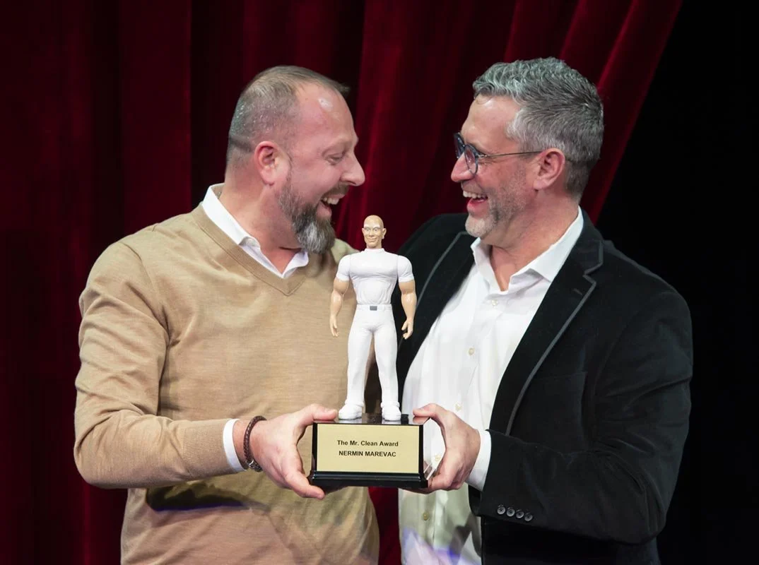 Two men smiling and holding an award with a small figurine on it, during an award ceremony with a red curtain backdrop.
