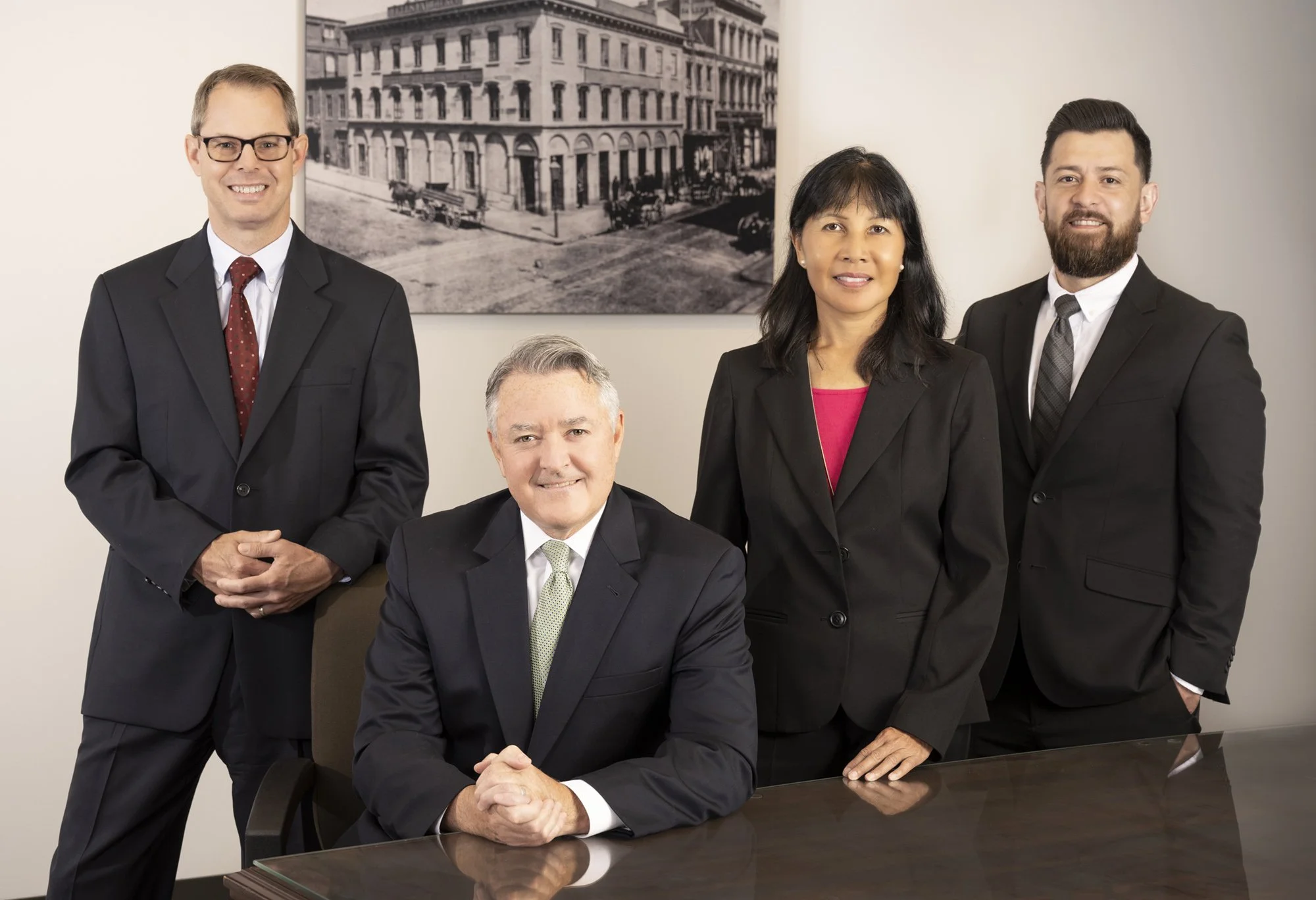 Four professionals in suits posing in an office, with a black and white cityscape photograph on the wall behind them. One man is seated at a wooden table, three are standing.