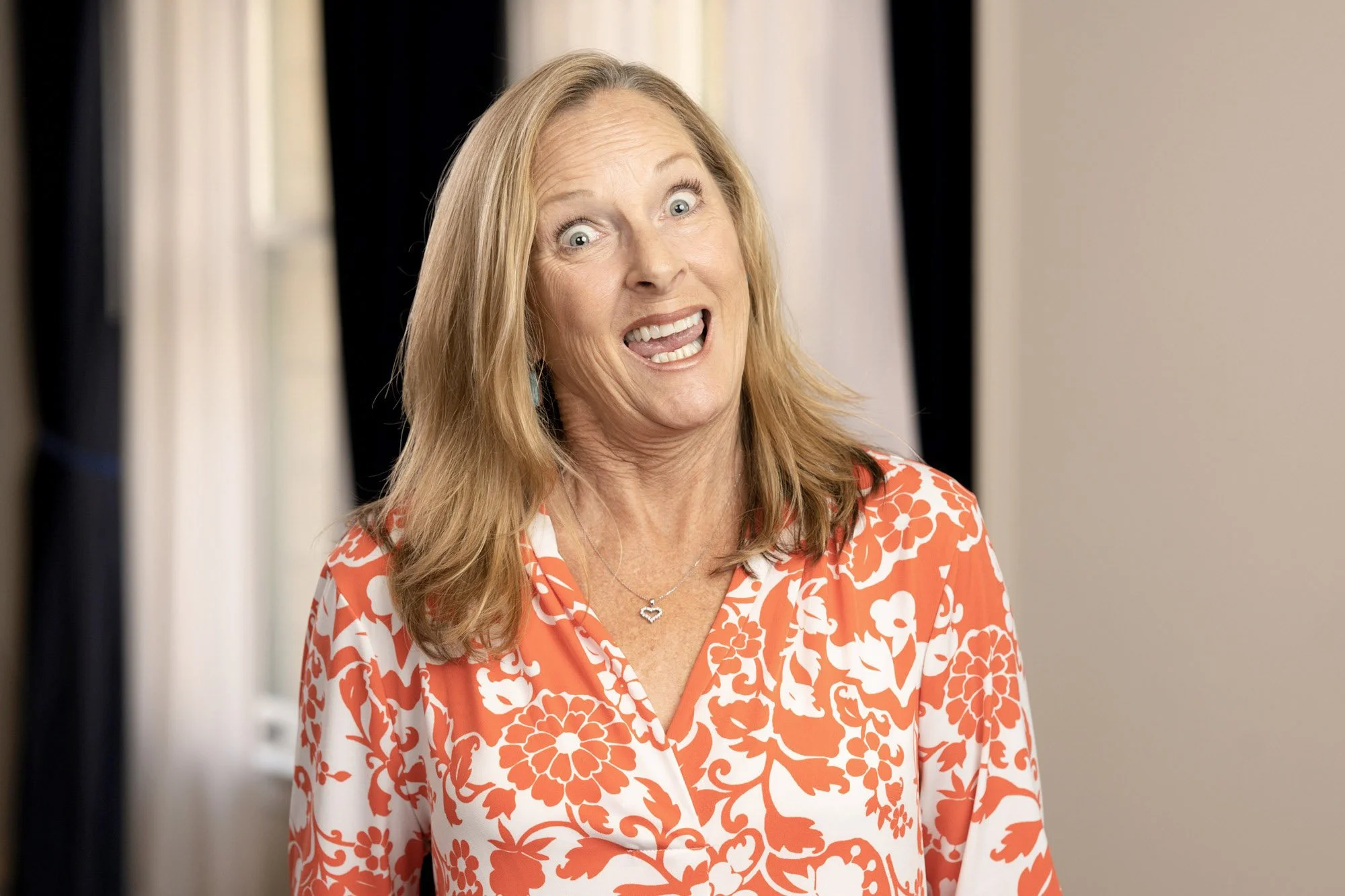 A woman with shoulder-length blonde hair, wearing a red and white floral blouse, has an excited or surprised expression with her mouth open and eyes wide as a blooper outtake.