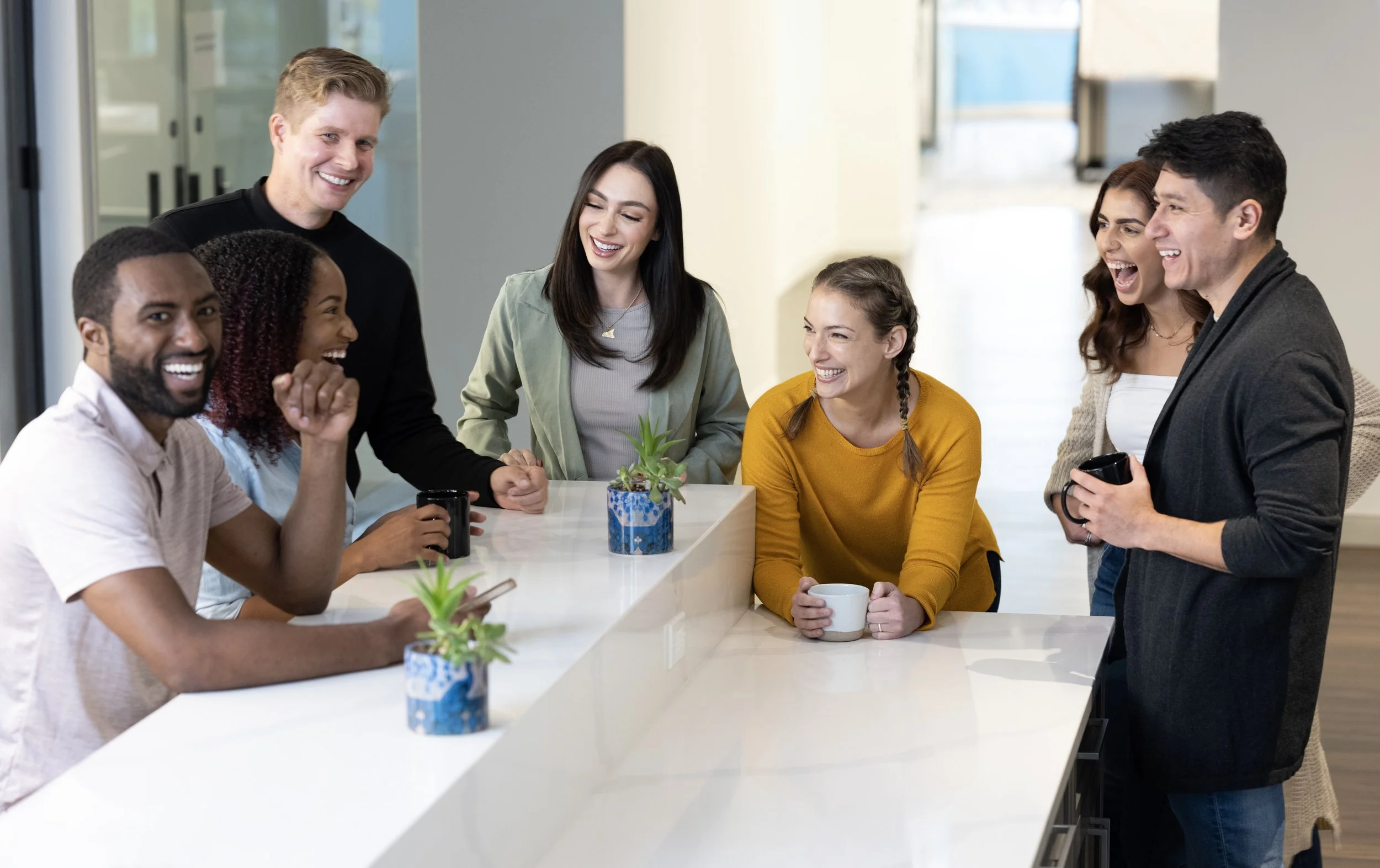 Group of six people laughing and talking around a white kitchen island, with some holding mugs, in a bright modern interior.