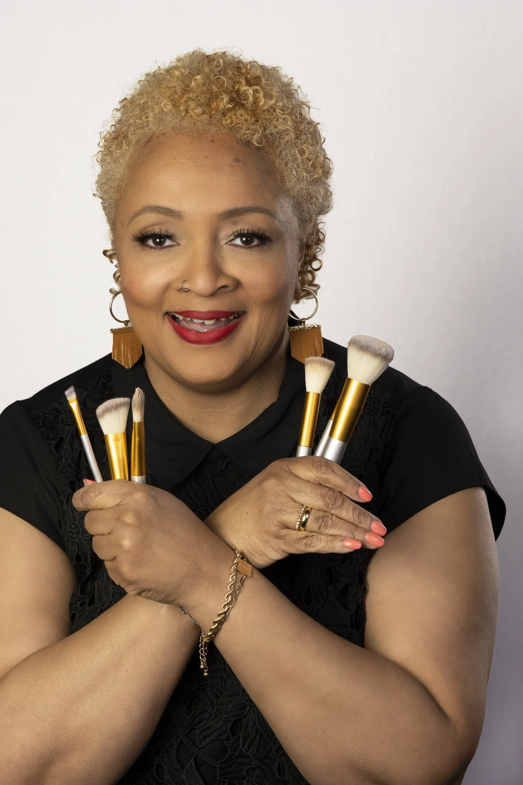 A makeup artist with short curly blonde hair holding makeup brushes and smiling to promote her business.