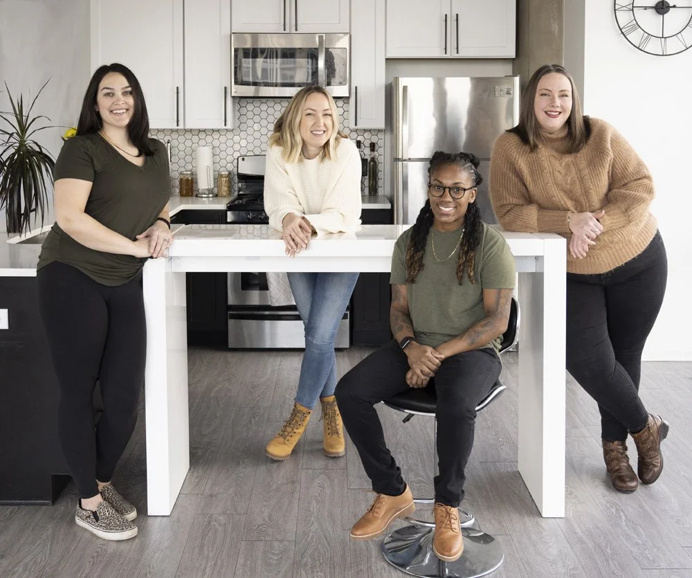 Five chefs smiling in a modern kitchen, some leaning on a white kitchen island and others sitting in chairs, with a stainless steel refrigerator and microwave behind them. A small personal chef Chicago business.