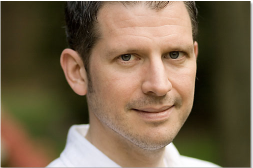 Close-up of a man with short dark hair, blue eyes, and light facial hair, wearing a white shirt, outdoors with blurred greenery in the background.