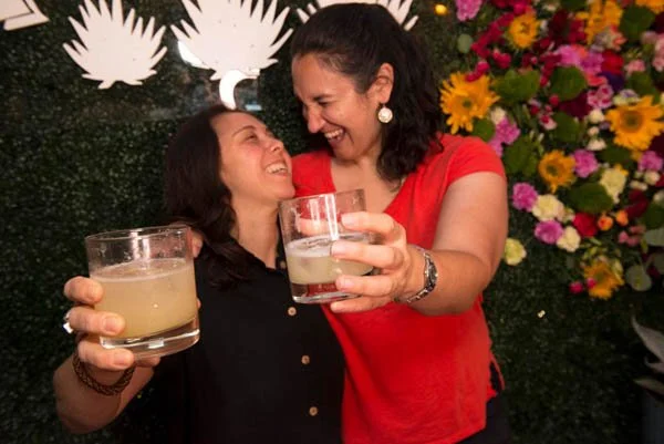 Two women smiling and holding drinks, celebrating together with a background of flowers and decorative clouds.