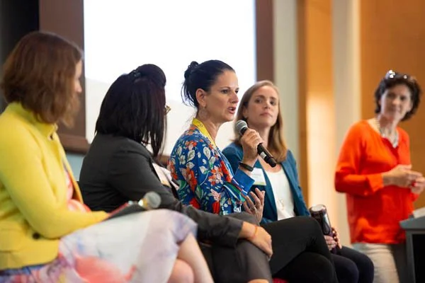Women sitting on a panel at a conference or seminar, with one woman speaking into a microphone.