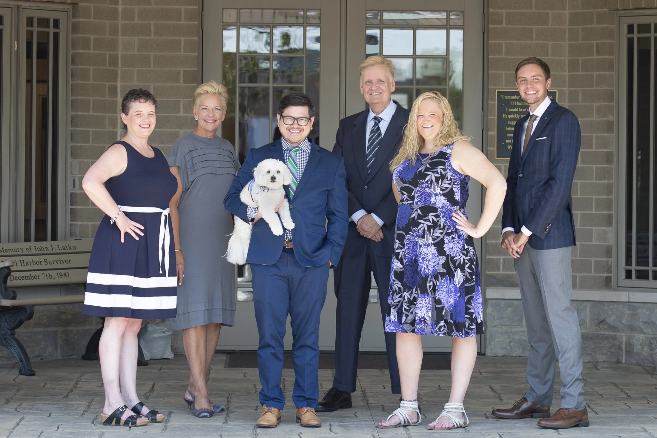 Group of seven people standing on a porch, smiling. One person is holding a small white dog. The background features a brick building and glass doors.