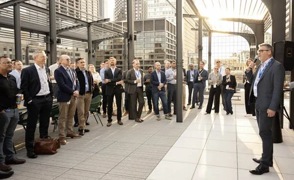 Group of business people listening to a speaker at an outdoor corporate event on a rooftop terrace with modern glass and steel architecture.