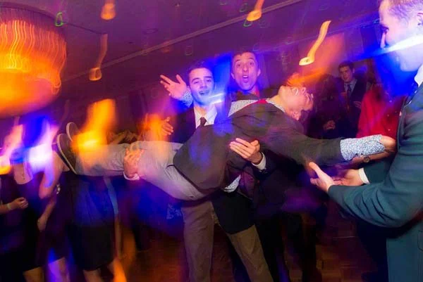 Group of people at a party holding a person horizontally, with colorful lighting and motion blur effects.