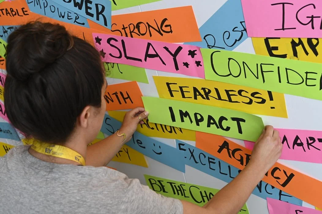 A person placing colorful strips of paper with words like 'SLAY,' 'FEARLESS!,' and 'IMPACT' on a wall covered with other handwritten words and phrases at a corporate event.