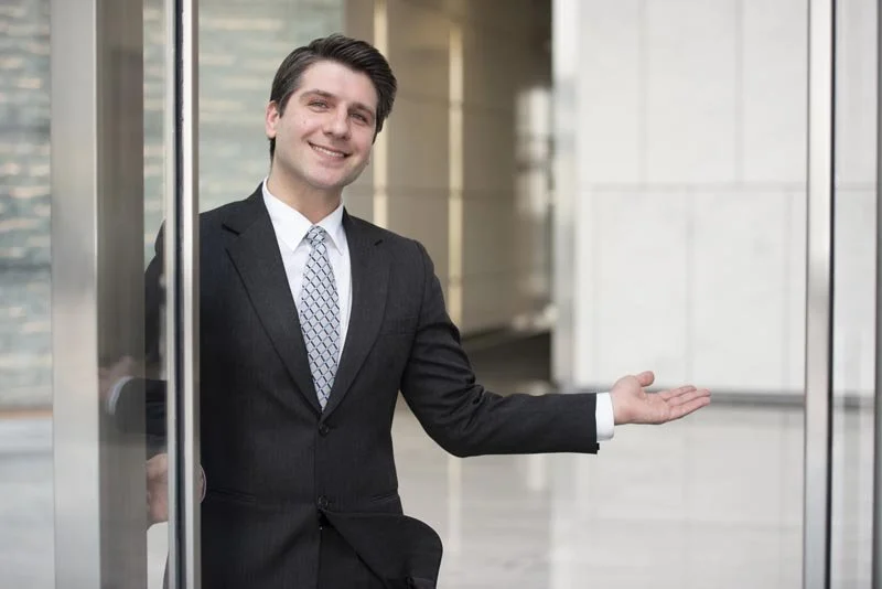 Businessman in a suit standing in an office building, gesturing with one hand.