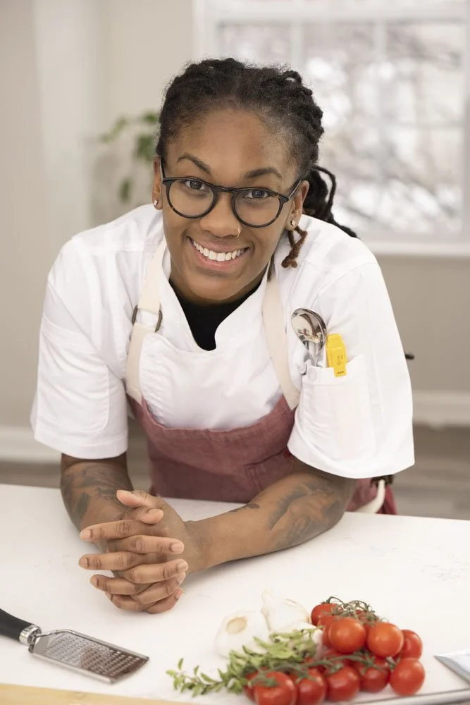 A smiling personal chef with glasses and dreadlocks, wearing a white chef's coat and pink apron, leaning over a kitchen counter with ingredients including tomatoes, garlic, and herbs, in a bright, homey kitchen. Promoting her small Chicago business.