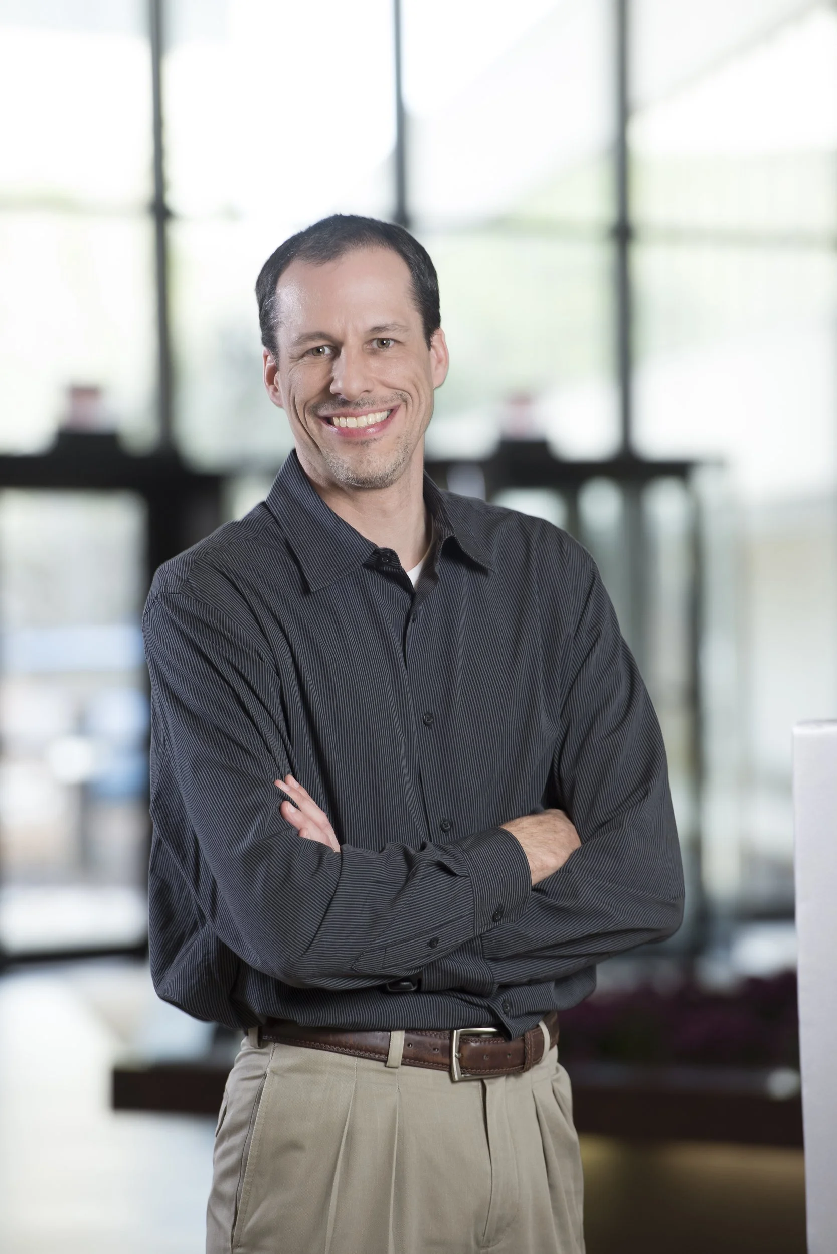 A smiling man with dark hair, wearing a dark gray button-up shirt and khaki pants, standing with arms crossed in front of a large window background.