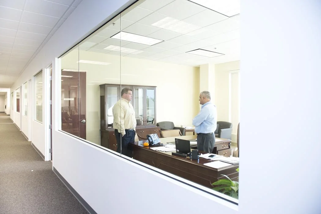 Two men having a conversation inside an office, viewed through a large glass window from a hallway.