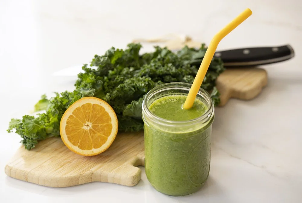 A glass jar filled with green smoothie, with a yellow straw, placed on a white surface as a bank of images for social media marketing. Behind, a halved orange, kale leaves, a knife, and a cutting board are visible.