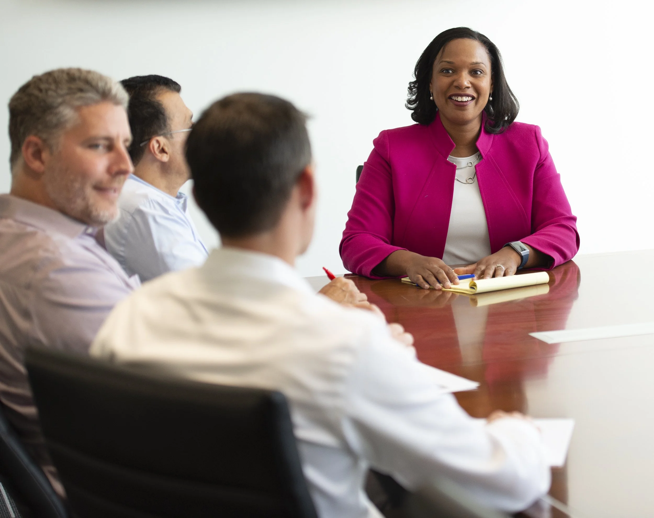 A woman in a bright pink blazer smiling during a business meeting with four people, seated around a wooden conference table.
