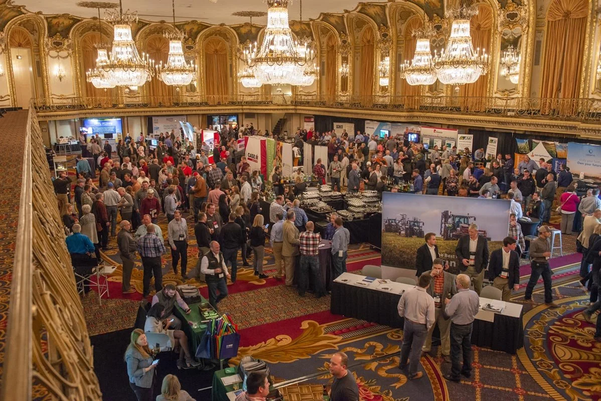 A large conference or trade show at the ornate ballroom at the Palmer House hotel filled with numerous booths and attendees walking around and talking. The room has elegant chandeliers, detailed gold trim, and a colorful patterned carpet.