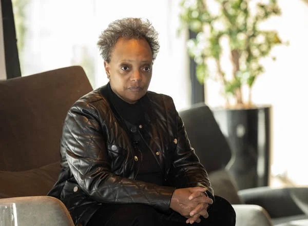 A woman with short, curly gray hair sitting on a brown chair indoors, wearing a black leather jacket and black clothing, with a somber expression and hands clasped.