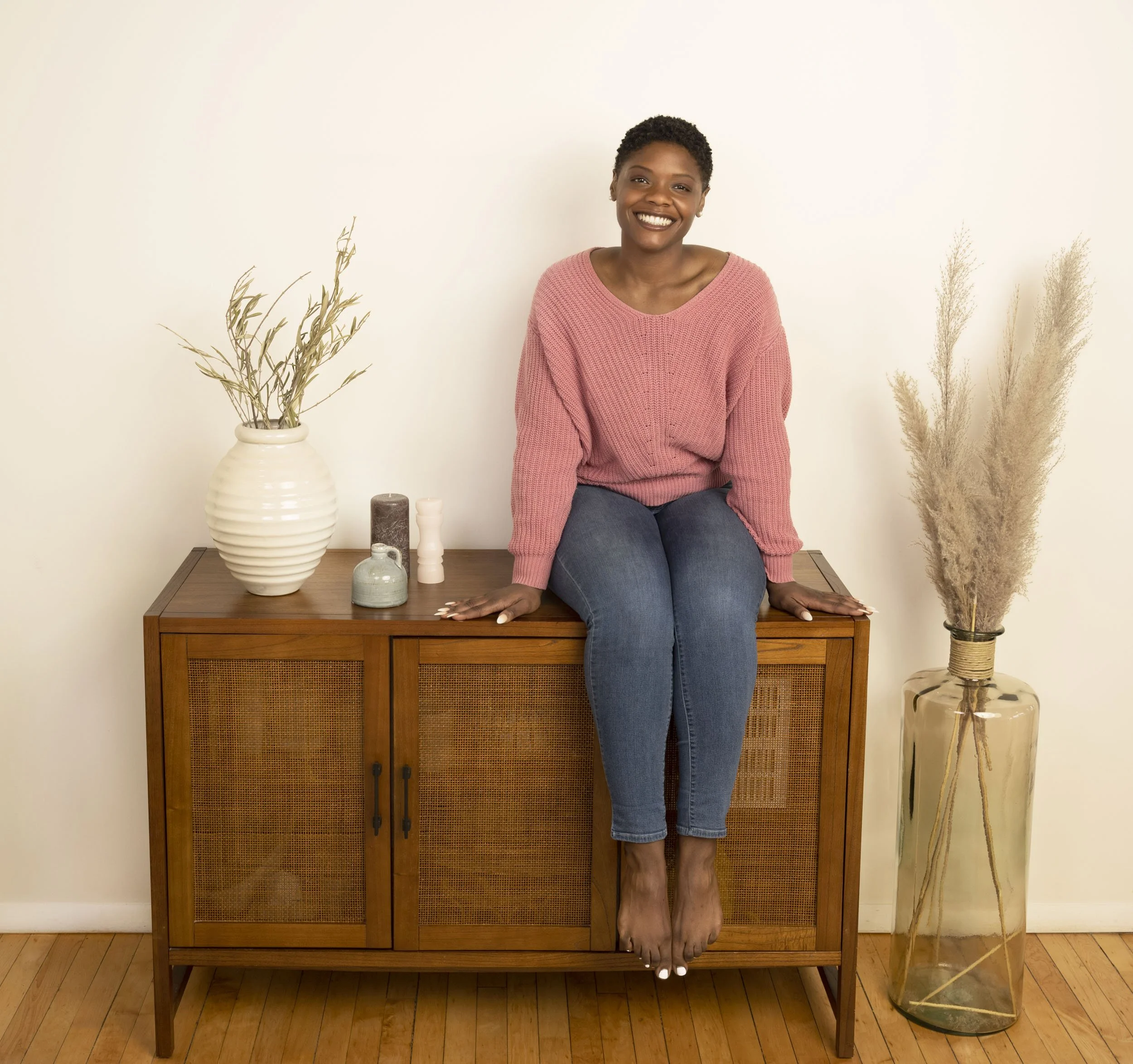 A black social media influencer, sitting on a wooden cabinet, wearing a pink sweater and blue jeans, smiling at the camera. There are decorative vases and dried plants on and around the cabinet.