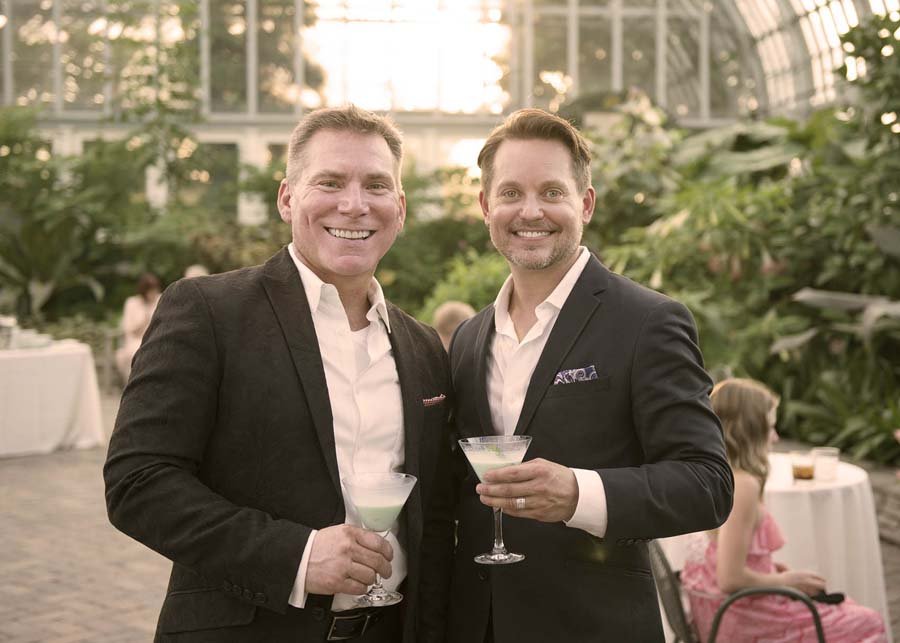 Two men in suits at a social event holding cocktails and smiling for the camera in a greenhouse or conservatory setting.