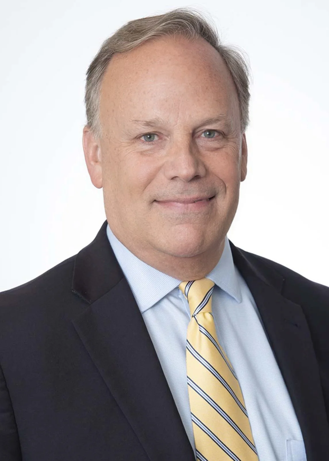 Professional headshot photography on white of a man in a dark blue suit and yellow tie smiling. The entire company was photographed on the same day.