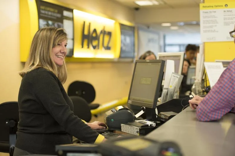 A woman with blonde hair smiling at a cashier at a Hertz rental car counter inside an airline terminal.